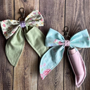 Könnte beinhalten: Zwei Stoff-Schleifen-Schlüsselanhänger mit Metallverschlüssen. Eine Schleife hat ein Blumenmuster mit grünen Akzenten, die andere ein blaues Blumenmuster mit einem rosa Lippenbalsamhalter. Beide befinden sich auf einem Holzgrund.