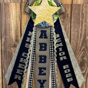 May include: A custom-made graduation sash with a gold glitter star, ribbons, and the name "ABBEY" in navy blue letters. The sash includes ribbons with the text "SENIOR 2024" and "HIGH SCHOOL". The design incorporates navy, silver, and black and white checkered patterns.