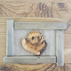 以下が含まれることがあります： 顔のような形をした自然な木彫りの木製ウォールアート。彫刻は長方形のフレーム内に配置されています。木材は明るい茶色で、濃いアクセントがあり、フレームは明るい灰色です。