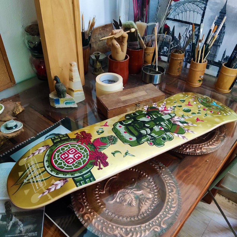 May include: A gold skateboard deck with a colorful graphic design featuring a green, blue, and red creature with a crown. The design also includes a red and white circle with the text "DEZA" inside. The skateboard deck is on a wooden surface.