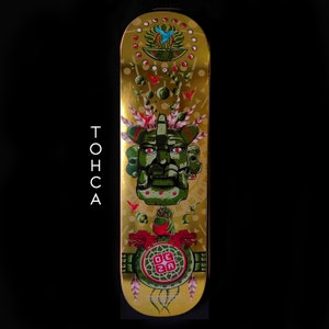 May include: Gold skateboard deck with a green and red illustration of a Mayan-style face with a headdress. The background is gold with red and blue birds and other geometric designs. The text "DEZA" is in red on a green circle.
