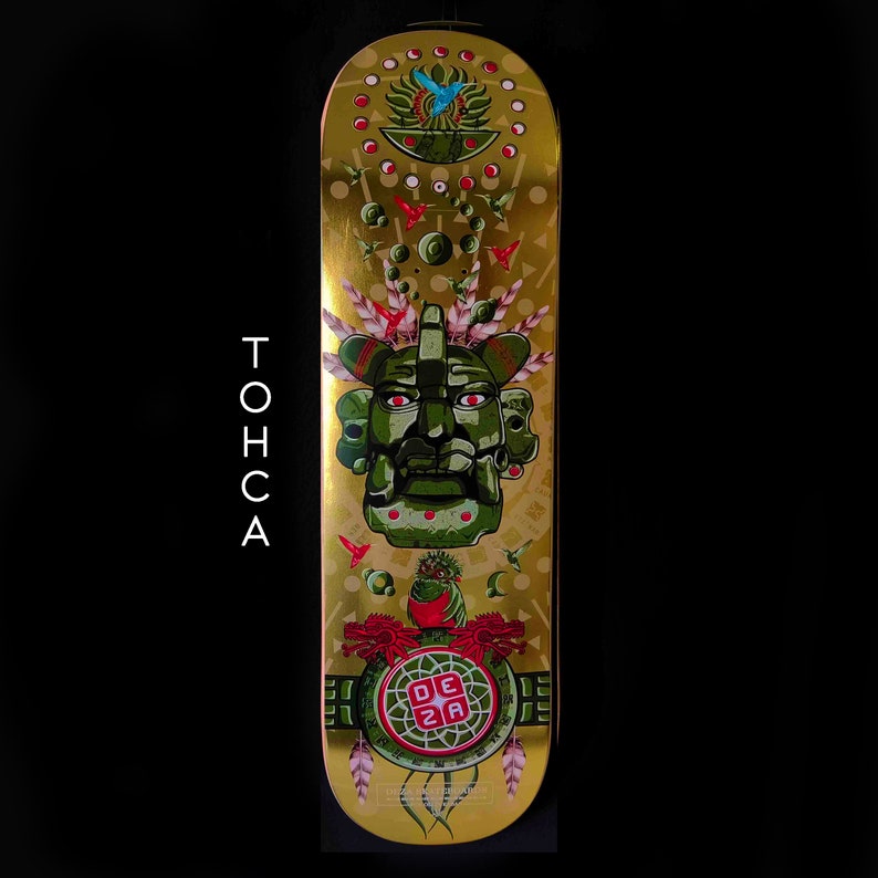 May include: Gold skateboard deck with a green and red illustration of a Mayan-style face with a headdress. The background is gold with red and blue birds and other geometric designs. The text "DEZA" is in red on a green circle.