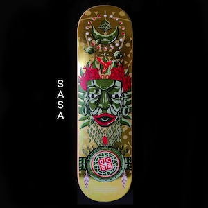 May include: A gold skateboard deck with a green and red design featuring a stylized face with a crown of red flowers. The text "DEZA" is in a red square on a green circle. The text "DIZA SKATEBOARDS" is at the bottom of the deck.
