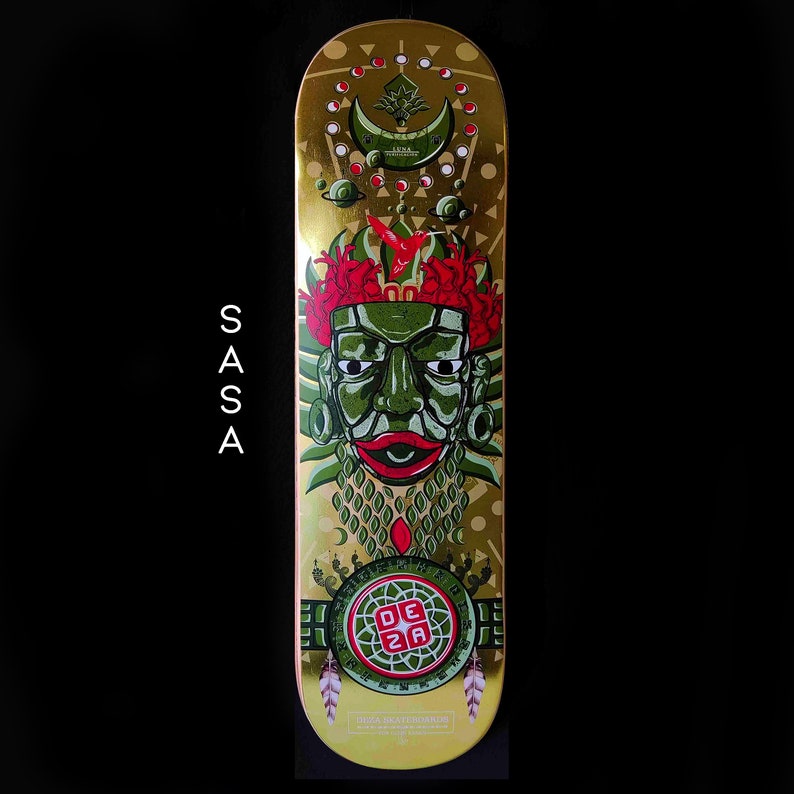 May include: A gold skateboard deck with a green and red design featuring a stylized face with a crown of red flowers. The text "DEZA" is in a red square on a green circle. The text "DIZA SKATEBOARDS" is at the bottom of the deck.