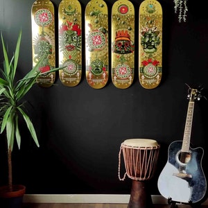 May include: Five gold skateboard decks with colorful graphic designs featuring mythical creatures and the text "DCZA" hanging on a black wall. A brown wooden drum and a black and white acoustic guitar are in the foreground.