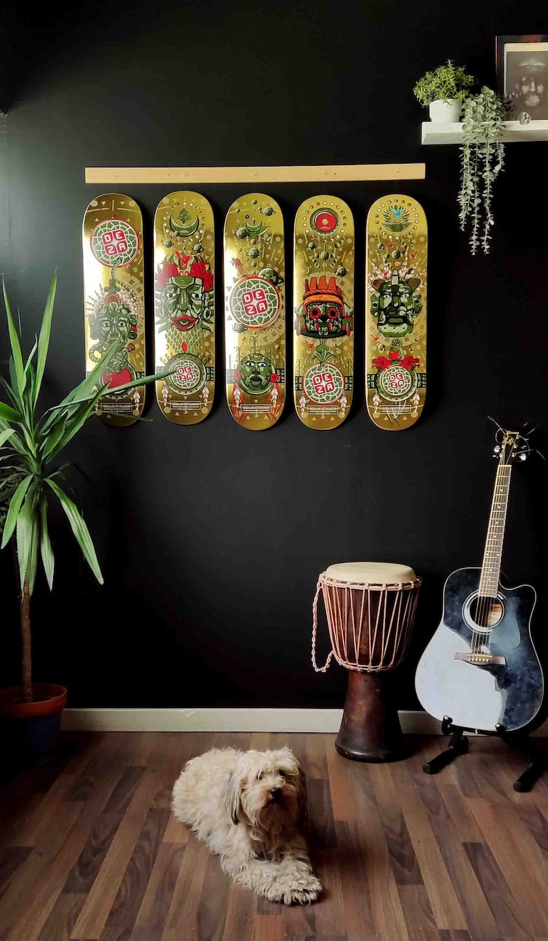 May include: Five gold skateboard decks with colorful graphic designs featuring mythical creatures and the text "DCZA" hanging on a black wall. A brown wooden drum and a black and white acoustic guitar are in the foreground.