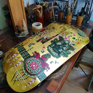 May include: Four gold-colored skateboards with colorful designs. The top skateboard features a green, red, and gold design with a green face and the text "DEZA SKATEBOARDS" and "MADE IN THE USA".