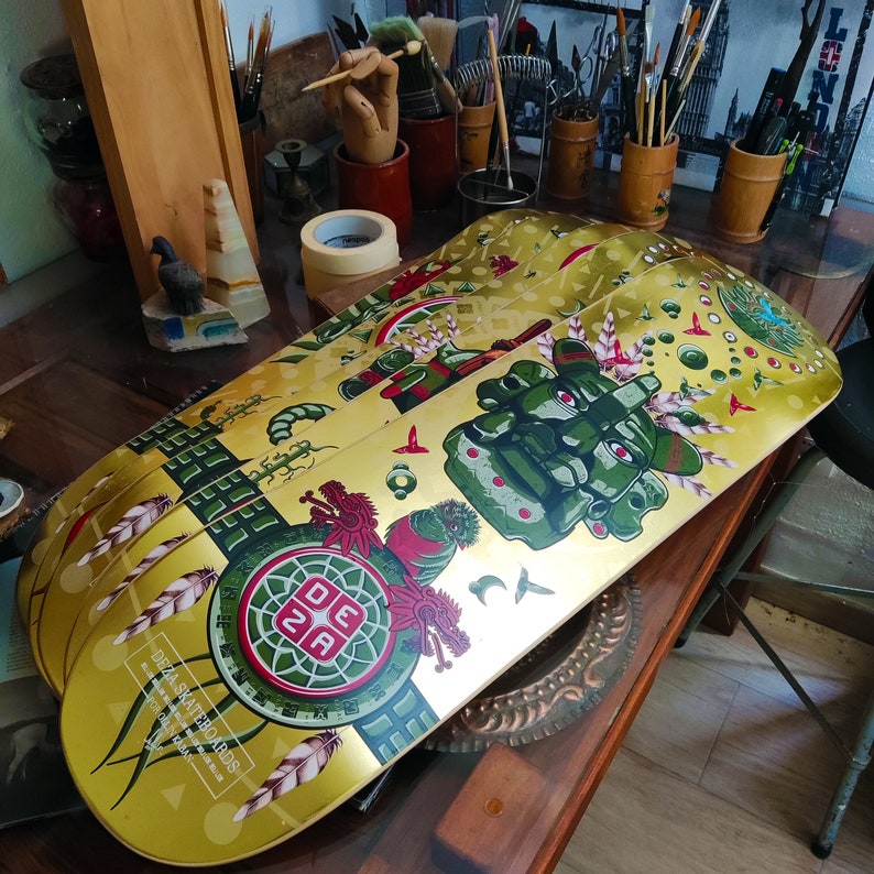 May include: Four gold-colored skateboards with colorful designs. The top skateboard features a green, red, and gold design with a green face and the text "DEZA SKATEBOARDS" and "MADE IN THE USA".