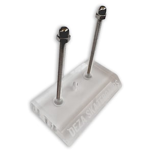 Puede incluir: Un soporte de exhibición de skateboard de acrílico transparente con dos tornillos de metal y una perilla plateada en cada tornillo. El soporte tiene las palabras "DEZA SKATEBOARDS" grabadas en la base.