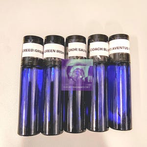 May include: Five blue glass roll-on bottles with black caps are displayed. The labels read "CREED: GREE", "GREEN IRISH", "DIOR: SAU", "COACH: BLU", and "AVENTUS T". The bottles are arranged vertically, suggesting a fragrance or essential oil product.