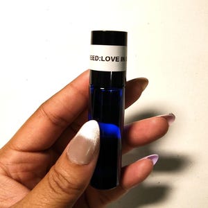 May include: A small, dark blue glass bottle with a black cap, held in a hand. The bottle has a white label with the text "NEED: LOVE IN". The bottle is filled with a dark blue liquid.