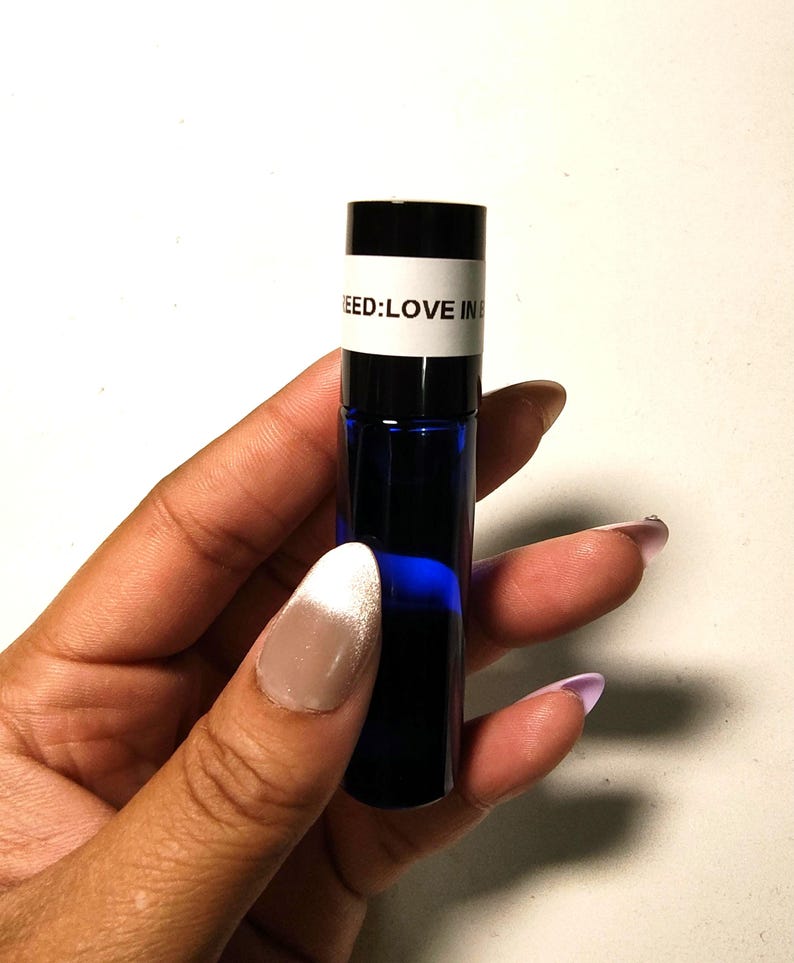 May include: A small, dark blue glass bottle with a black cap, held in a hand. The bottle has a white label with the text "NEED: LOVE IN". The bottle is filled with a dark blue liquid.