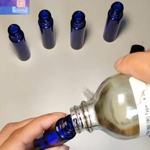 May include: Several small, cobalt blue glass bottles with dark blue screw-on caps. A clear glass bottle is being poured into one of the blue bottles. The bottles are on a light-colored surface.