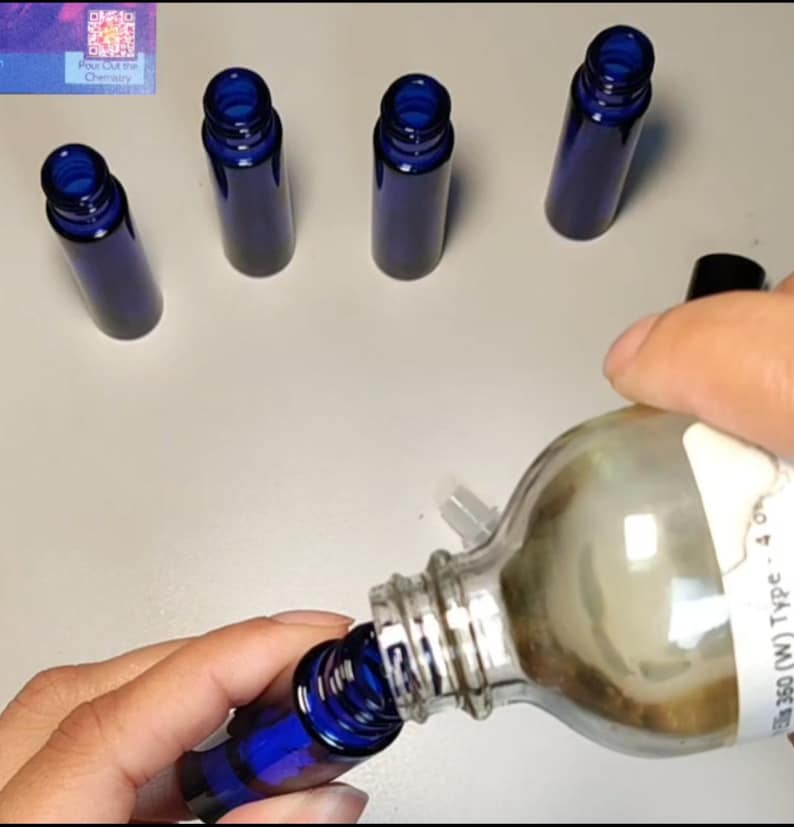 May include: Several small, cobalt blue glass bottles with dark blue screw-on caps. A clear glass bottle is being poured into one of the blue bottles. The bottles are on a light-colored surface.