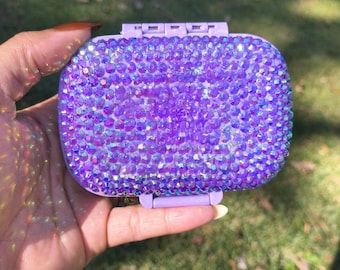 May include: A rectangular, lilac compact mirror encrusted with iridescent purple rhinestones. The compact has a hinged lid and a small clasp. The background is blurred green grass.