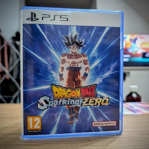 Peut inclure: Boîtier de jeu PS5 pour Dragon Ball Sparking! ZERO. La couverture présente un personnage musclé aux cheveux noirs hérissés, vêtu de vêtements orange et bleus, sur fond d'explosion d'énergie bleue. Le jeu est classé 12 et comprend le logo Bandai Namco.