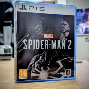 Puede incluir: Una caja de juego de PlayStation 5 azul con una imagen en blanco y negro de Spider-Man en la parte delantera. El juego está clasificado para mayores de 16 años y se titula "Marvel's Spider-Man 2".