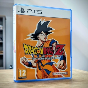 May include: A blue PlayStation 5 game case with an orange background. The case features a cartoon image of Goku from Dragon Ball Z, with the game title "Dragon Ball Z: Kakarot" printed in white. The game is rated 12+ by PEGI and is published by Bandai Namco.