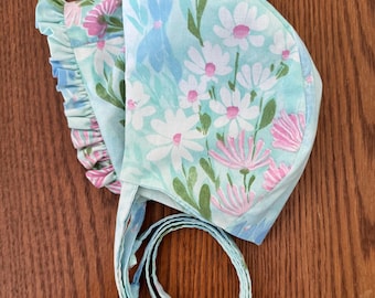 May include: A baby bonnet with a floral pattern in shades of blue, white, pink, and green. The bonnet features a ruffled edge and ties. The fabric is decorated with daisies, bunny silhouettes, and other floral elements.