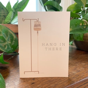 May include: A beige card with a brown IV bag hanging from a brown metal stand. The text "HANG IN THERE" is printed in brown letters on the card.
