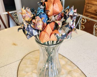 Puede incluir: Un jarrón de cristal transparente contiene un ramo de flores de papel de origami. Las flores son de varios colores, incluyendo naranja, azul y diseños estampados. El jarrón está sobre una mesa blanca con un patrón decorativo similar al mármol.