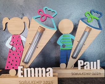 Puede incluir: Decoraciones de cono escolar de madera con figuras de niña y niño, cada una con un tubo de vidrio. La niña lleva un vestido rosa, y el niño una camisa a rayas azules y verdes. Se muestran los nombres "Emma" y "Paul", junto con "SCHULKIND 2025".