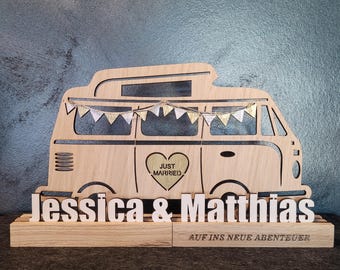 May include: Wooden wedding cake topper featuring a camper van design with the words "Just Married" inside a heart. The names "Jessica & Matthias" are displayed below the van, with "AUF INS NEUE ABENTEUER" underneath.