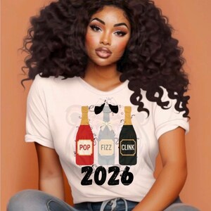 Pode incluir: Camiseta branca com um gráfico de três garrafas de champanhe com as palavras "POP", "FIZZ" e "CLINK". O número "2026" está impresso abaixo. A imagem mostra uma mulher com cabelo encaracolado escuro.