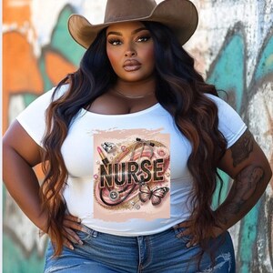 Puede incluir: Camiseta blanca con un dise&ntilde;o gr&aacute;fico que presenta un estetoscopio, l&aacute;piz labial, diamantes y una mariposa. El gr&aacute;fico tambi&eacute;n incluye la palabra "NURSE" en letras doradas con purpurina.