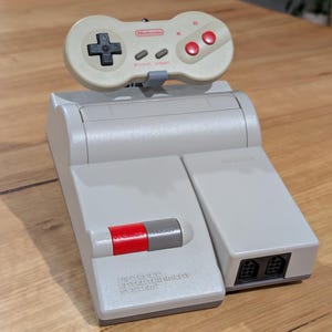May include: A grey Nintendo Entertainment System console with a matching controller. The controller is off-white with red and grey buttons. The console has a red and grey power button and the words "Nintendo Entertainment System" printed on the front.