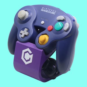 Peut inclure: Une manette Nintendo GameCube violette avec un support violet assorti. La manette comporte divers boutons colorés et un joystick blanc. Le support présente le logo GameCube. La manette est connectée à un câble noir.