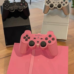 Może przedstawiać: Trzy konsole i kontrolery Sony PlayStation 2 w kolorze czarnym, srebrnym i różowym. Kontrolery są umieszczone na konsolach. Różowa konsola i kontroler są na pierwszym planie, a za nimi srebrna i czarna konsola.