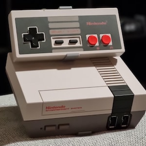 Op de afbeelding: Een klassieke Nintendo Entertainment System console met bijbehorende controller. De console is lichtgrijs met een donkergrijze streep en zwarte accenten. De controller heeft een kruis, knoppen en rode start/select-knoppen. Het Nintendo-logo is zichtbaar.
