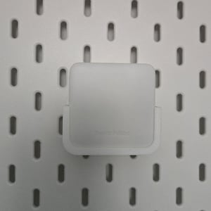 May include: A white SwitchBot device mounted on a white pegboard. The device is square with rounded edges and the text "SwitchBot" is visible. The pegboard has evenly spaced oval holes.