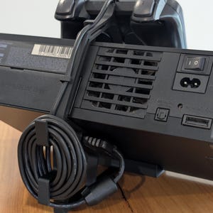 May include: A black video game console with a matching controller and power cord. The console has a power button and ventilation slots. The controller is positioned on top of the console, and the power cord is neatly coiled and secured.