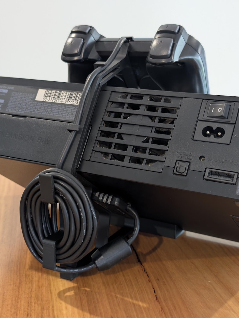 May include: A black video game console with a matching controller and power cord. The console has a power button and ventilation slots. The controller is positioned on top of the console, and the power cord is neatly coiled and secured.