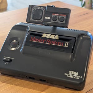 May include: A black Sega Master System II video game console with a detached controller. The console has the Sega logo and "Master System II" printed on the front. The controller has a directional pad and two buttons. The console is on a wooden surface.