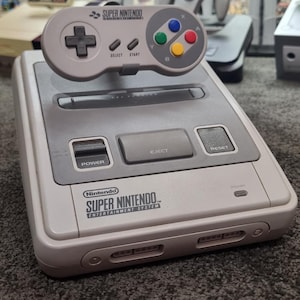 May include: A light grey Super Nintendo Entertainment System console with a matching controller. The console has grey buttons and the Super Nintendo logo. The controller features a directional pad, coloured buttons, and the words "SELECT" and "START".