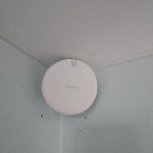 May include: A white, circular Aqara smart home device fixed to a pale blue wall. The device has a small notch at the top and the brand name is printed in a light grey font. The device is installed in the corner of a room.
