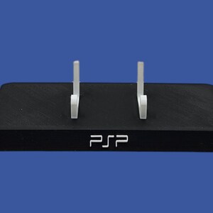 Pu&ograve; includere: Supporto rettangolare nero con due bracci di supporto bianchi. Il supporto ha le lettere "PSP" in bianco sul davanti. Lo sfondo &egrave; blu unito.