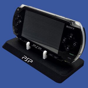 Pu&ograve; includere: Console di gioco portatile Sony PSP nera in una custodia protettiva trasparente, appoggiata su un supporto nero con le lettere bianche "PSP". La console ha uno schermo nero e pulsanti di controllo.