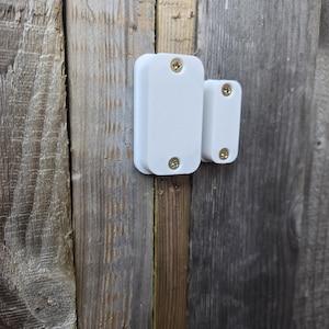 May include: Two white rectangular security sensors with rounded edges are fixed to a weathered wooden surface. Each sensor is secured with two gold-coloured screws. The wood has a mix of grey and brown tones.