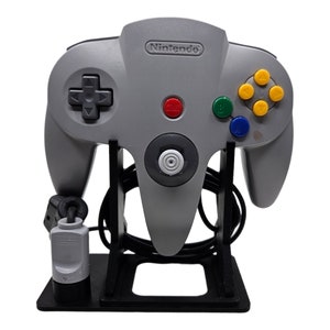 Op de afbeelding: Een grijze Nintendo 64-controller met een zwart kruis, een rode startknop en kleurrijke actieknoppen. De controller wordt weergegeven op een zwarte standaard, met de kabel en stekker zichtbaar. Het Nintendo-logo staat op de controller.