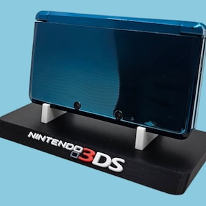 Puede incluir: Una consola de juegos portátil Nintendo 3DS de color turquesa sobre un soporte negro con soportes blancos. El soporte presenta el logotipo de Nintendo 3DS en blanco y rojo. El fondo es azul claro.