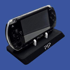 Pu&ograve; includere: Una console di gioco portatile Sony PSP nera appoggiata su un supporto nero. Il supporto presenta le lettere bianche "PSP". La PSP ha una finitura argentata e pulsanti neri. Lo sfondo &egrave; blu.