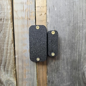 May include: Two black rectangular metal plates with rounded corners, textured surfaces, and gold-coloured rivets. The plates are mounted on a weathered wooden background, with one plate slightly larger than the other.