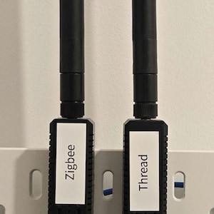 Può includere: Due dispositivi neri Zigbee e Thread con antenne montati su una pannello forato bianco. Ogni dispositivo ha un'etichetta bianca con il nome della tecnologia. Cavi blu e bianchi collegati.