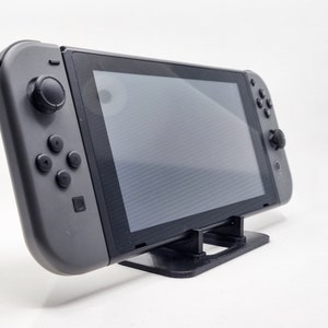 Może przedstawiać: Szara konsola do gier przenośna z czarnym ekranem jest oparta na czarnym stojaku. Konsola ma przyciski i joysticki po obu stronach. Stojak jest prostokątny z dwoma pionowymi wspornikami.