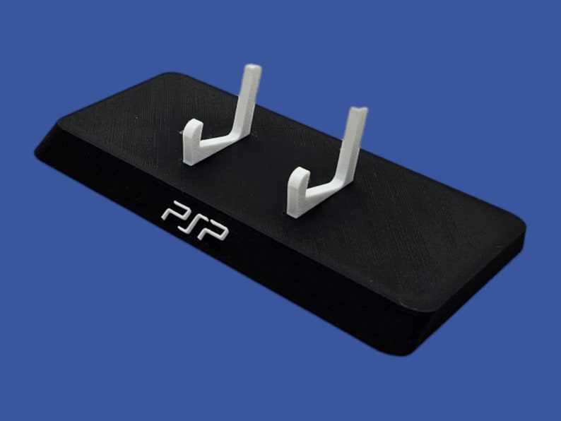 Pu&ograve; includere: Un supporto nero per console PSP con due bracci di supporto bianchi. Il supporto ha le lettere "PSP" in bianco sul davanti. Il supporto &egrave; rettangolare con un bordo anteriore leggermente angolato.