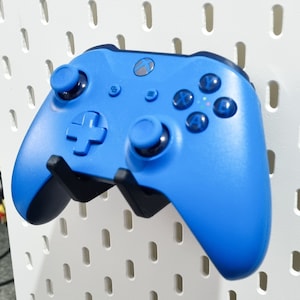 May include: A vibrant blue Xbox controller with black detailing is displayed on a white pegboard. The controller features a directional pad, buttons, and joysticks. The Xbox logo is visible.